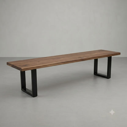 A long wooden bench with a rustic brown wood top and black metal U-shaped legs, photographed in a minimalist studio setting with a gray floor and wall.