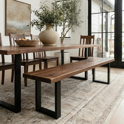 A dining room scene featuring a wooden table with black metal legs, a matching wooden bench, and dark wood chairs, styled with a large beige vase of greenery on a patterned rug.