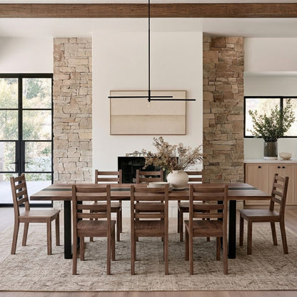 A modern dining room featuring a dark wood rectangular table and matching wooden chairs, styled with a white vase of dried branches on top, set on a textured beige rug in a contemporary home interior with stone columns and a fireplace.