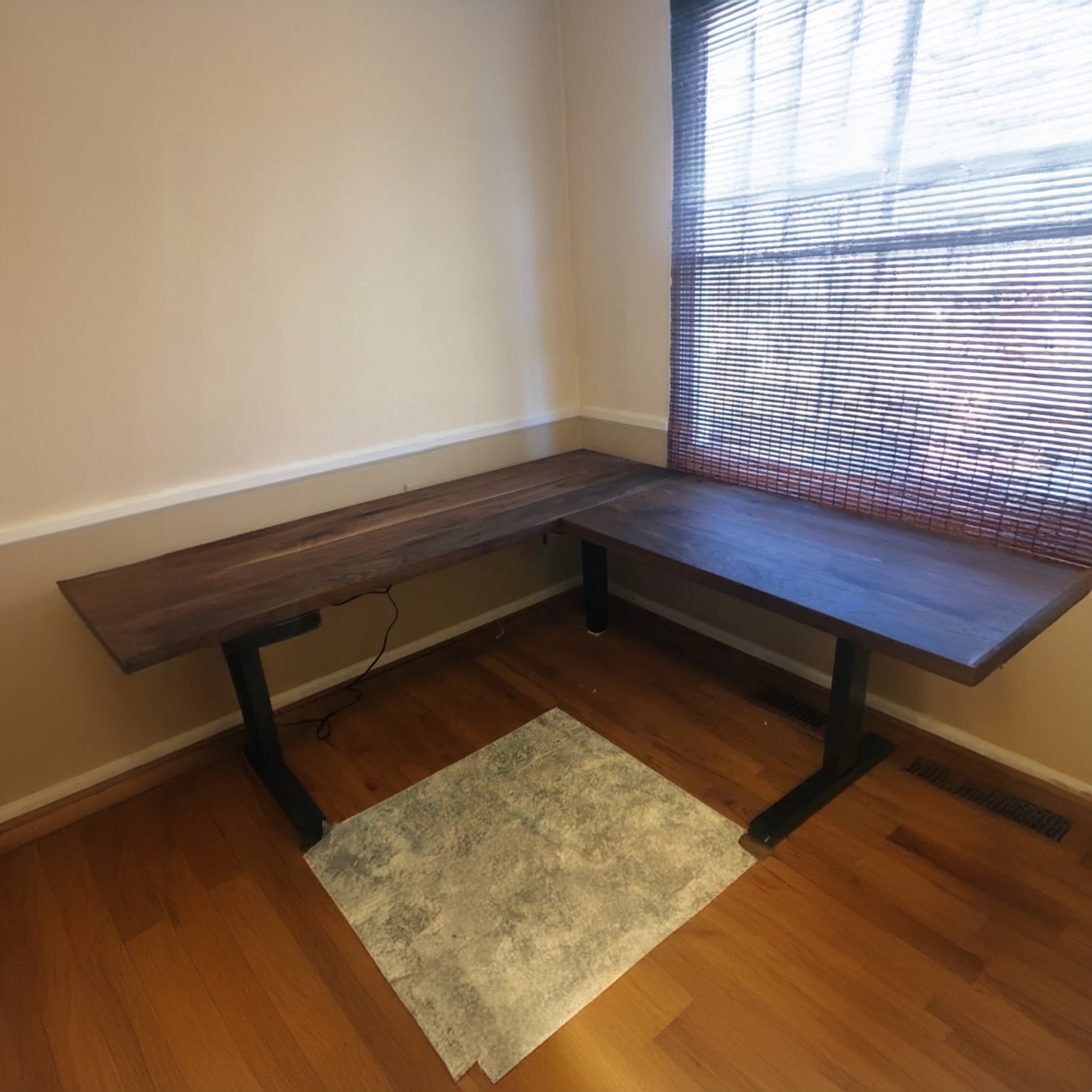 An L-shaped walnut desk with modern black metal legs, placed in an office setting with a chair and window in the background.