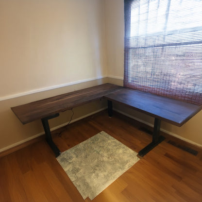 An L-shaped walnut desk with modern black metal legs, placed in an office setting with a chair and window in the background.