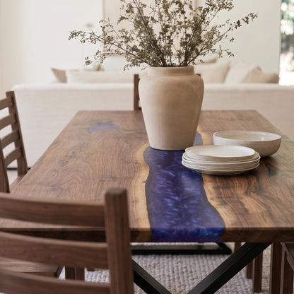 A rustic wooden dining table with a glossy dark blue resin inlay, styled with a large beige ceramic vase of dried branches, a stack of white plates, and a bowl in a bright, modern dining room.