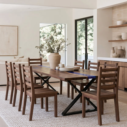 A modern dining room set featuring a rectangular wooden table with a blue inlay and black X-frame base, surrounded by matching wooden ladder-back chairs, set on a textured light rug in a bright, contemporary dining room.