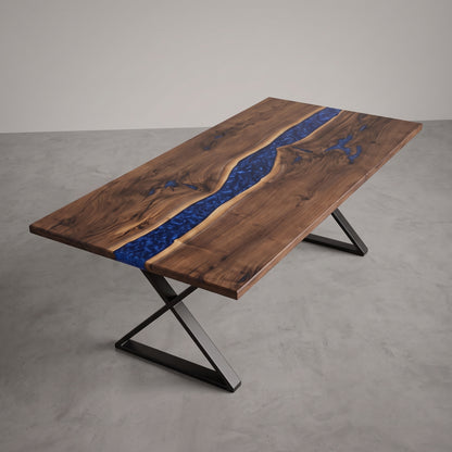 A rectangular wooden table with a live-edge design, featuring a vibrant blue resin river inlay running down the center. The table is supported by a black metal X-frame base and is photographed in a minimalist studio setting with a gray concrete floor and neutral gray wall.