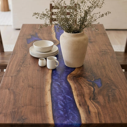 A rustic wooden dining table with a live edge and a prominent dark blue resin river running down the center, styled with a beige ceramic vase of dried flowers, stacked white ceramic dishes, and two small white mugs.