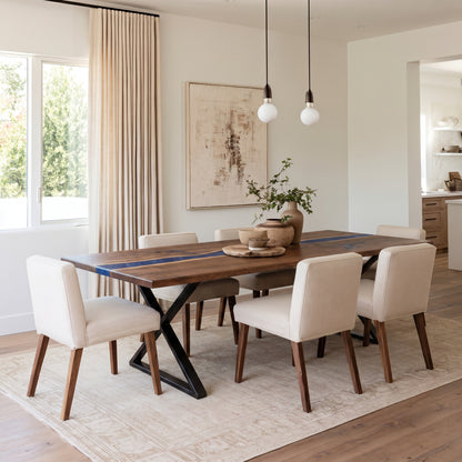 A modern dining room set featuring a rectangular wooden table with a blue runner and black X-base, surrounded by six cream upholstered dining chairs on a light beige rug.
